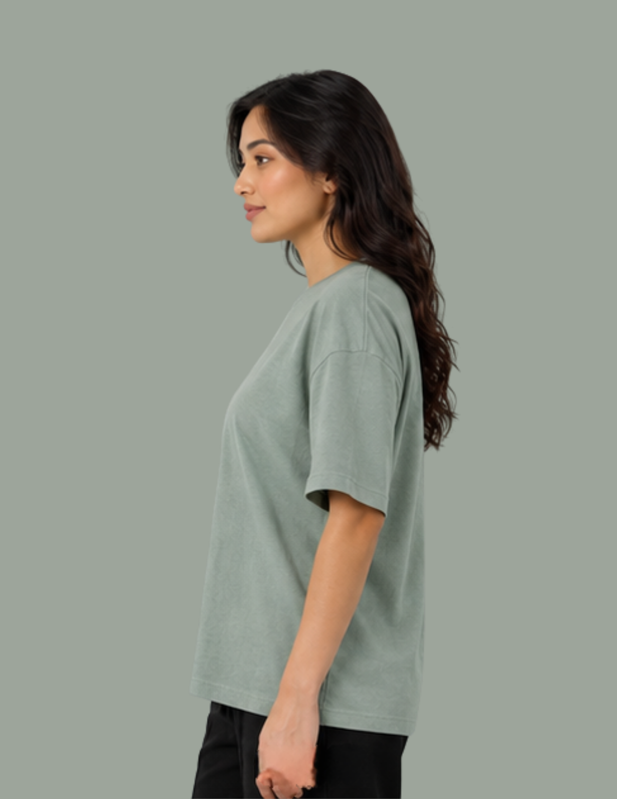 A woman shown in side profile wearing an oversized short-sleeve crewneck t-shirt in a muted sage green color, paired with black pants, against a solid sage green studio background.