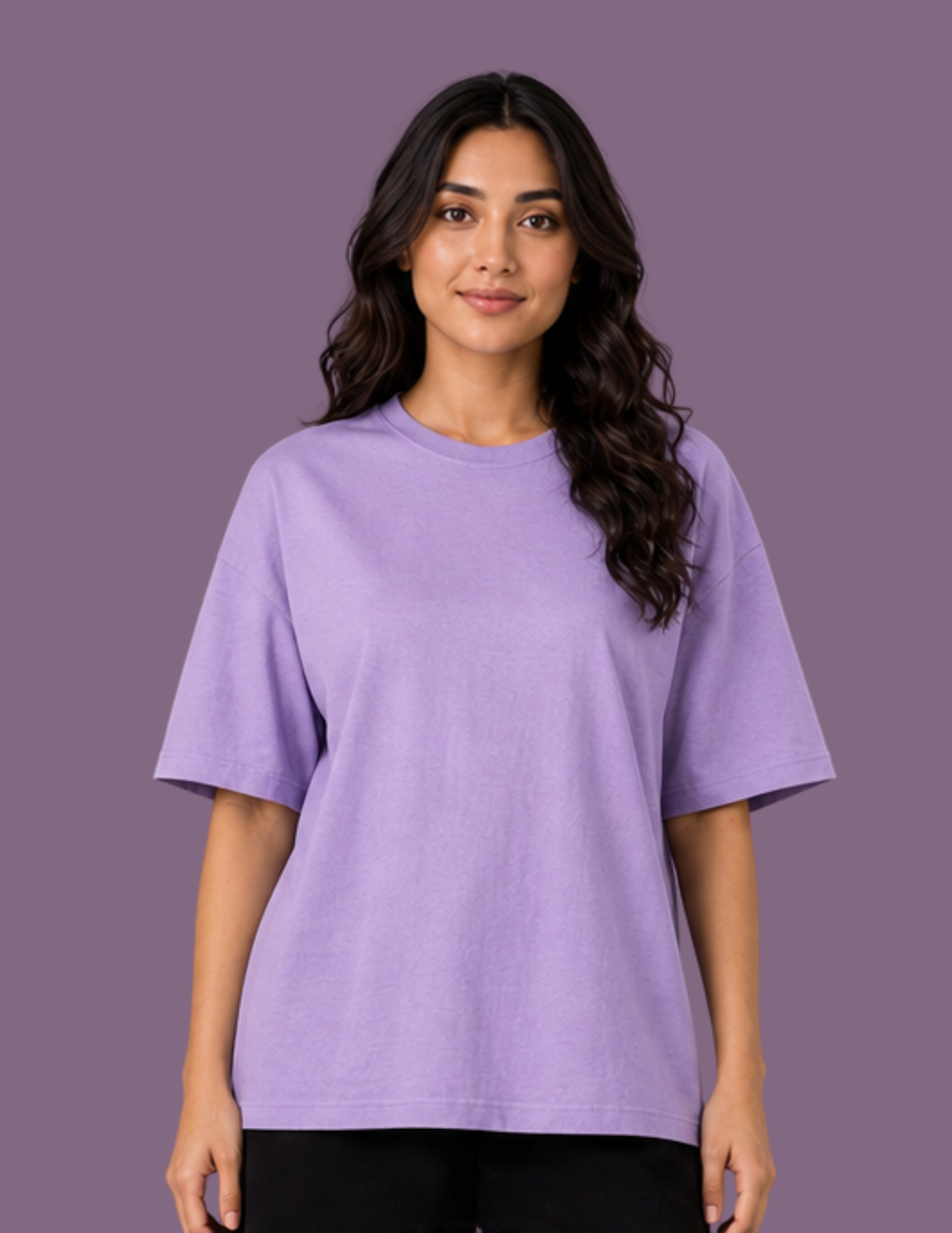A woman with long, wavy dark hair wearing an oversized short-sleeve crewneck t-shirt in light purple, paired with black pants, standing against a solid purple studio background.