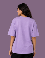 A woman viewed from the back wearing an oversized lavender-purple t-shirt and black pants, set against a solid purple studio background.