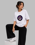 A woman models a white oversized t-shirt featuring a colorful cosmic eye graphic on the chest, paired with black wide-leg pants and white sneakers, posed on a white block against a solid gray background.