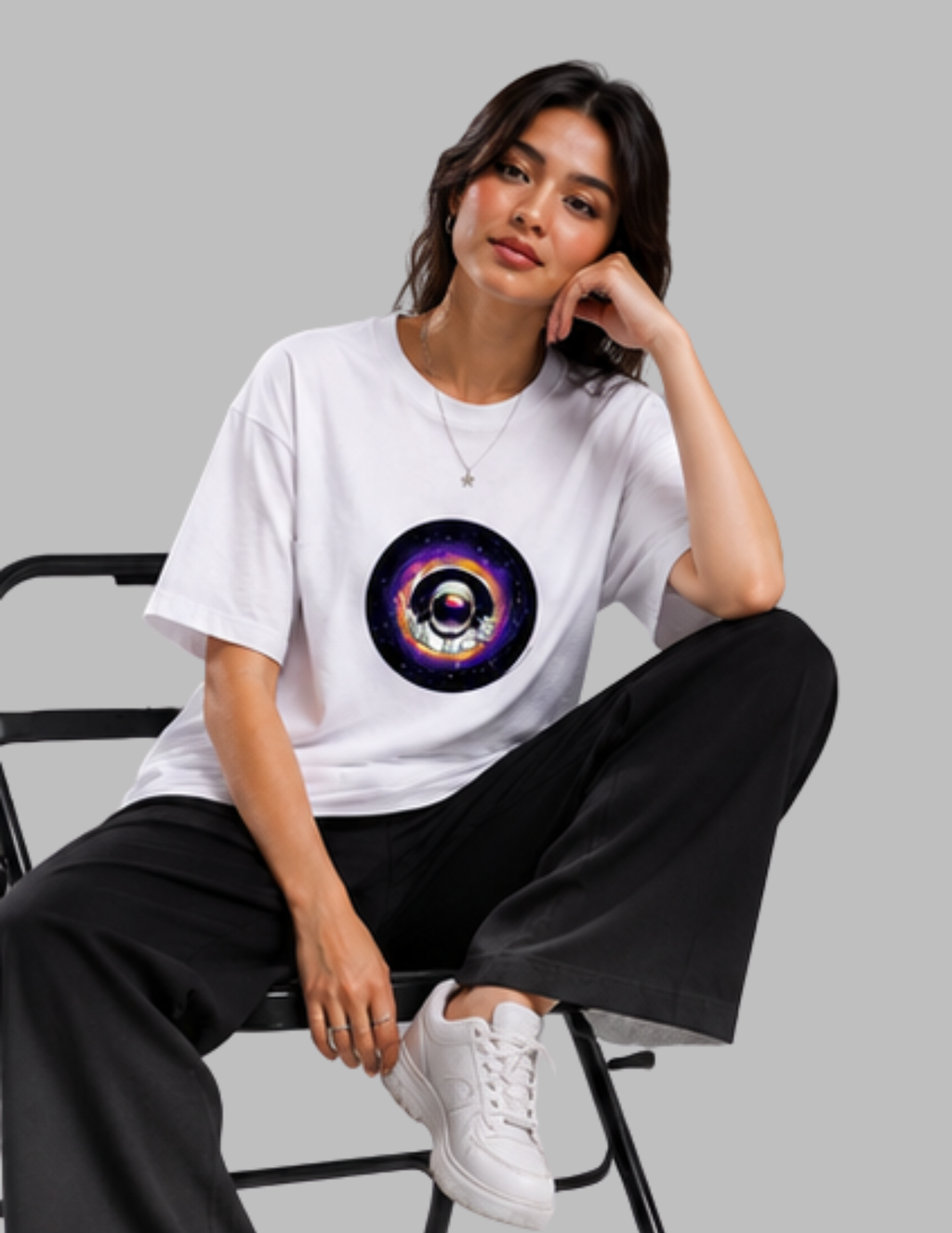 A woman sitting on a black chair wearing a white crewneck t-shirt with a colorful cosmic eye graphic on the chest, paired with black wide-leg pants and white sneakers, against a solid gray background.