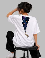 A woman wearing a white oversized T-shirt with a large back graphic of a space-themed bat silhouette and astronaut, seated on a black stool against a light gray background.