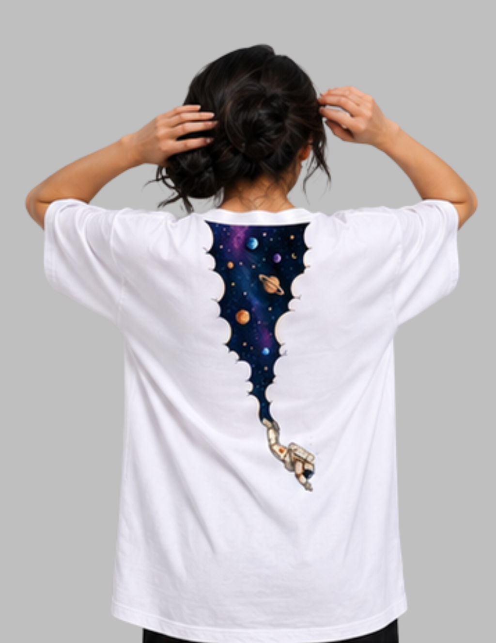 A person viewed from behind wearing a white short-sleeve T-shirt with a large space-themed graphic on the back. The design shows a torn-paper effect revealing a colorful galaxy scene with planets and a small astronaut.
