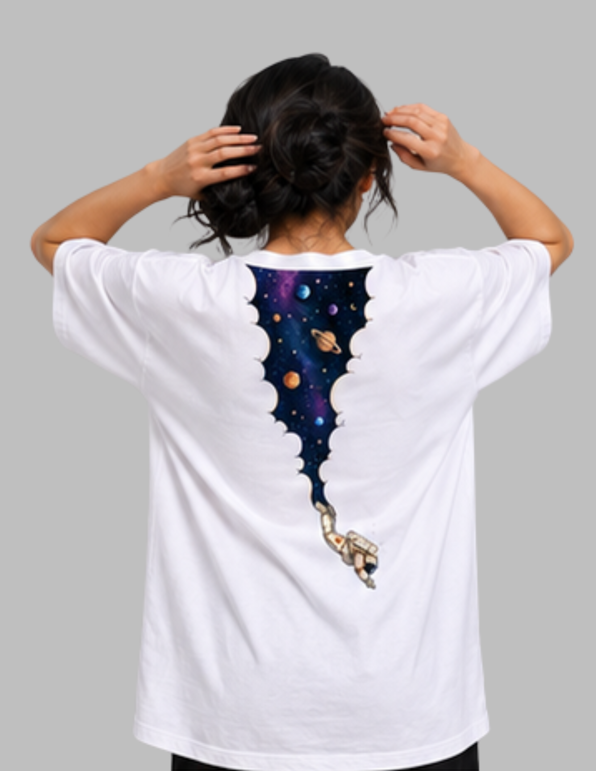 A person viewed from behind wearing a white short-sleeve T-shirt with a large space-themed graphic on the back. The design shows a torn-paper effect revealing a colorful galaxy scene with planets and a small astronaut.