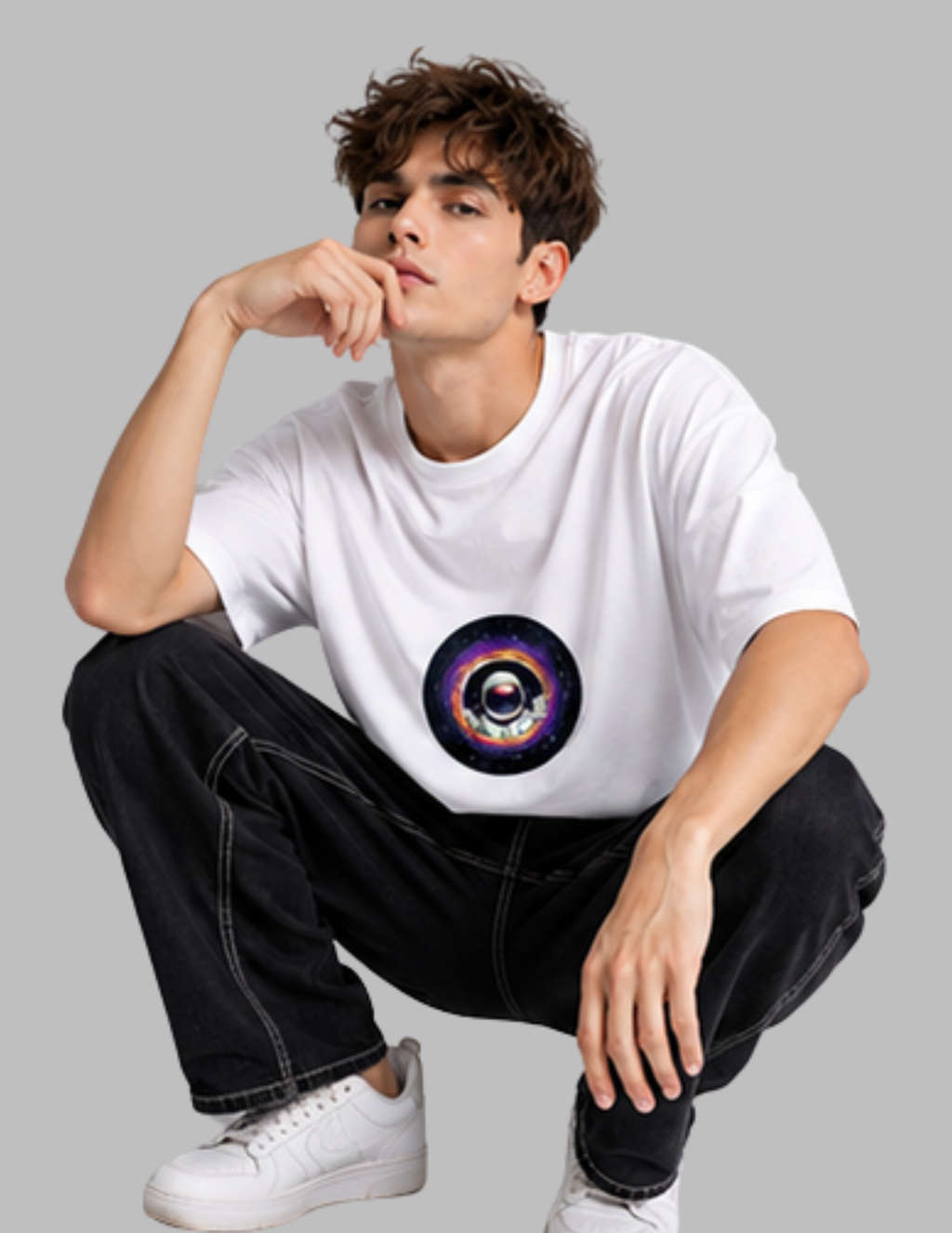 A young man wearing a white crewneck t-shirt with a colorful cosmic eye graphic on the chest, paired with black jeans and white sneakers, posing against a solid gray background.