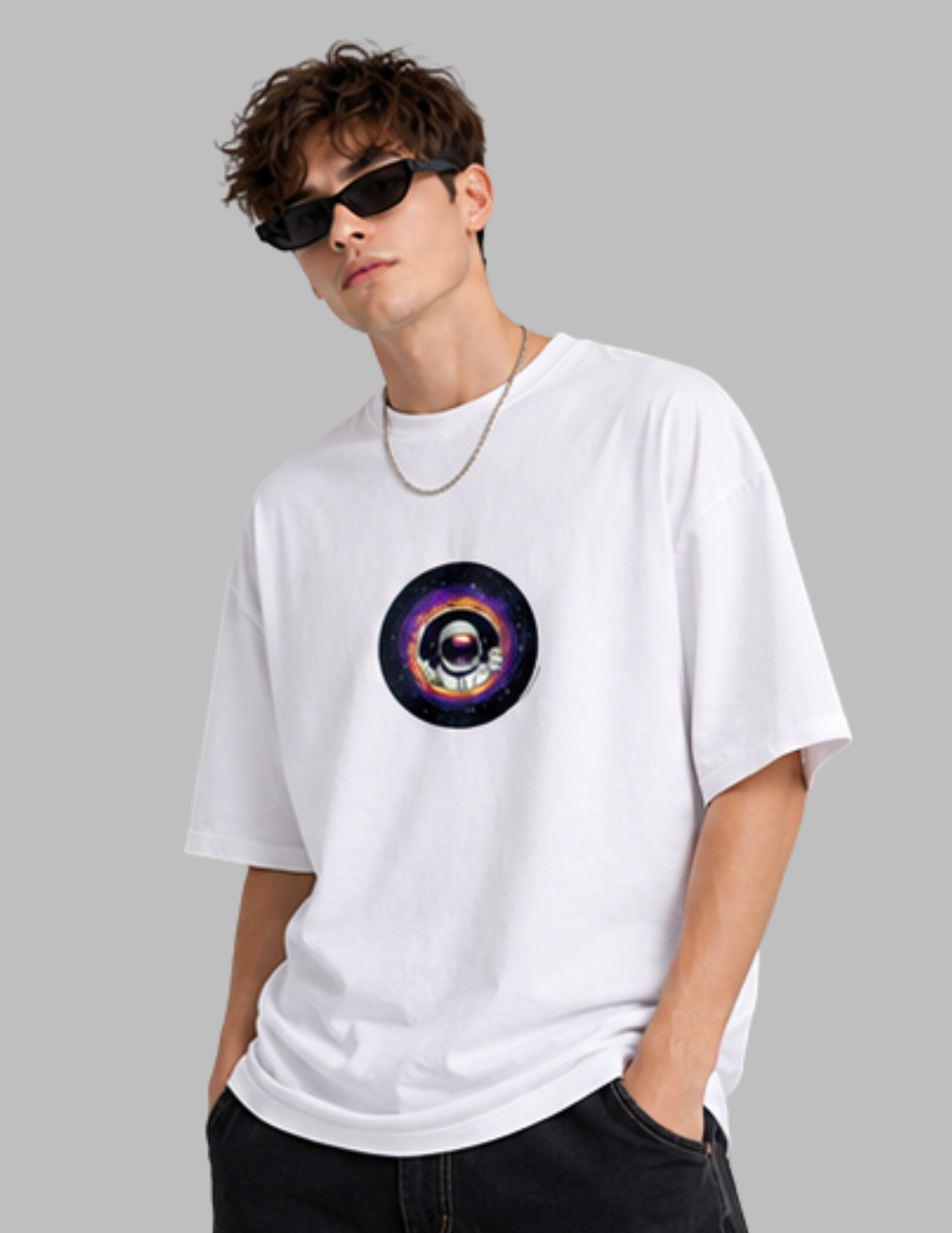 A man wearing a white oversized t-shirt with a colorful cosmic eye graphic on the chest, styled with black sunglasses, a silver chain necklace, and black jeans against a solid gray background.