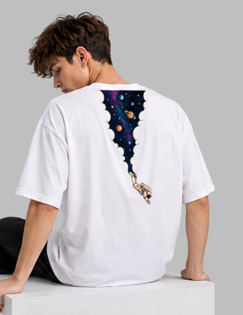 A man wearing a white oversized t-shirt, viewed from the back. The shirt features a graphic print of a cosmic space scene with planets and stars inside a cloud-like arch, with a small astronaut floating below.