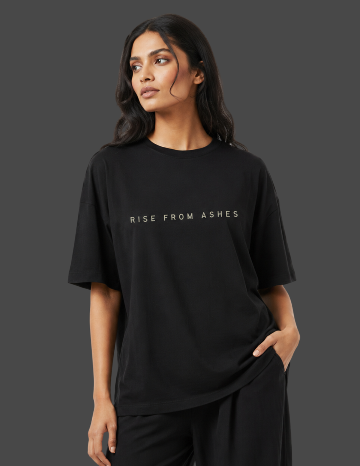 A woman wearing a black oversized short-sleeve t-shirt with the text 'RISE FROM ASHES' printed in white across the chest, paired with black pants, set against a solid dark gray background.