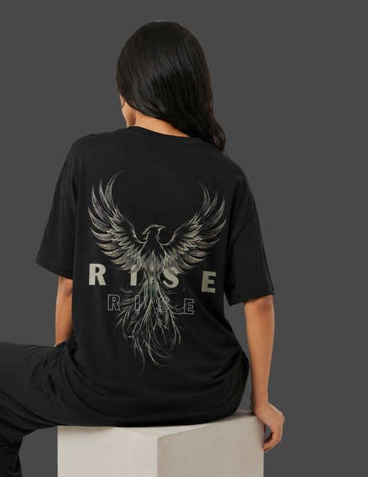 A woman shown from behind wearing a black oversized T-shirt with a large metallic silver phoenix graphic and the word 'RISE' printed across the back, seated on a light gray block against a dark gray background.