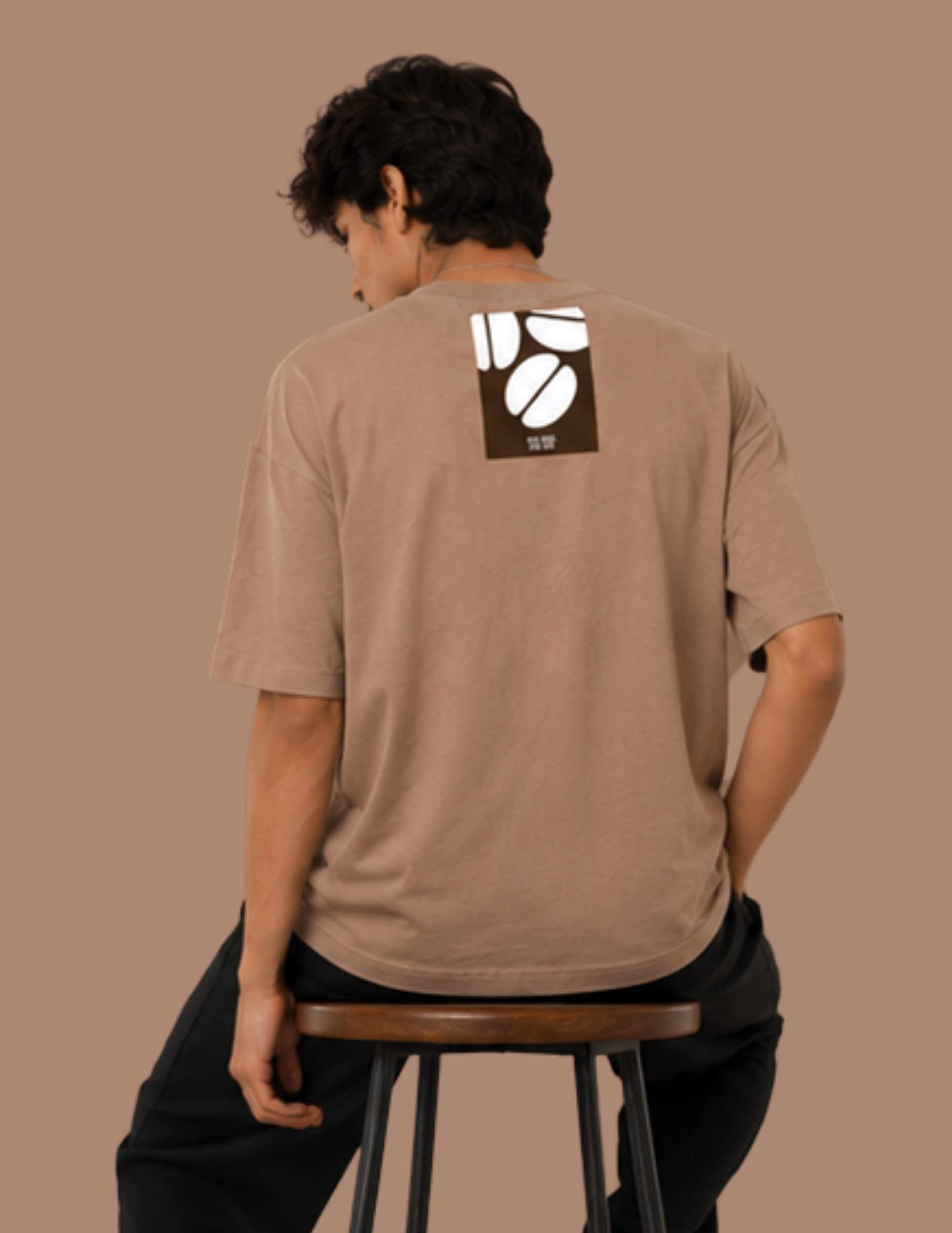 A man wearing a brown t-shirt with a coffee bean graphic on the back, sitting on a wooden stool against a solid brown background.