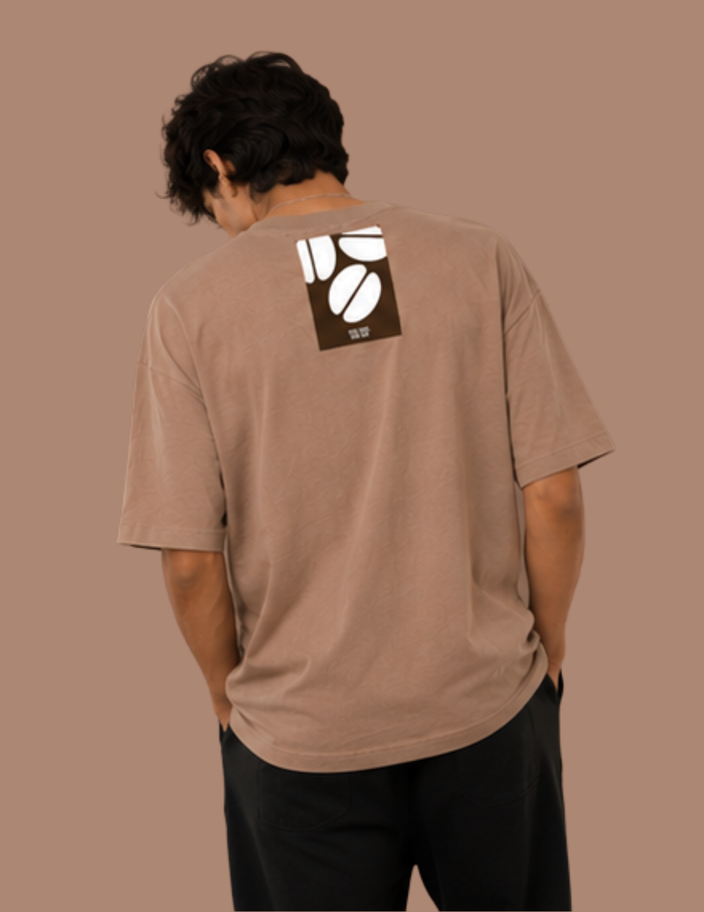 A man viewed from behind wearing a brown t-shirt with a white and dark brown coffee bean graphic on the upper back, paired with black pants, set against a solid brown background.