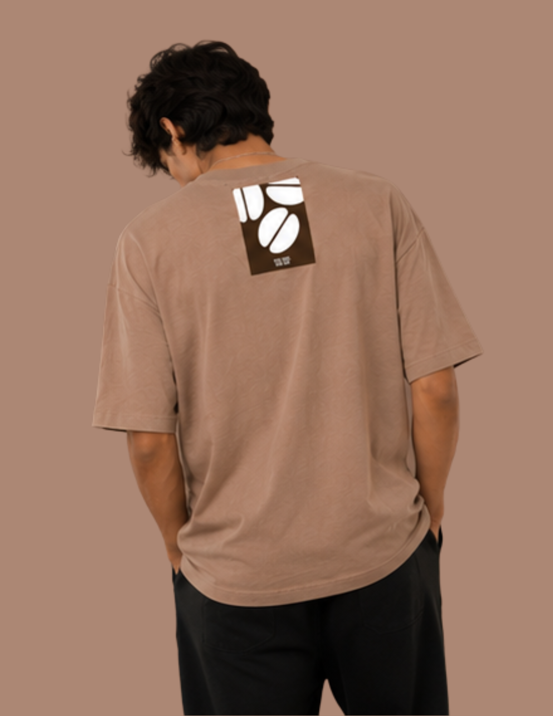 A man viewed from behind wearing a brown t-shirt with a white and dark brown coffee bean graphic on the upper back, paired with black pants, set against a solid brown background.