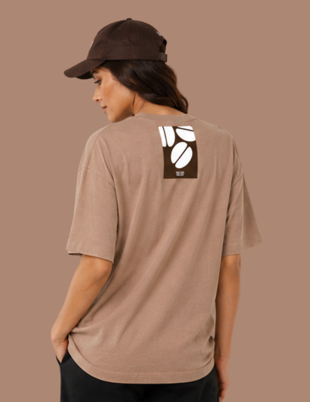 A woman wearing a brown baseball cap and a taupe-brown oversized t-shirt, shown from the back. The t-shirt features a square graphic on the upper back with white coffee beans on a dark brown background.