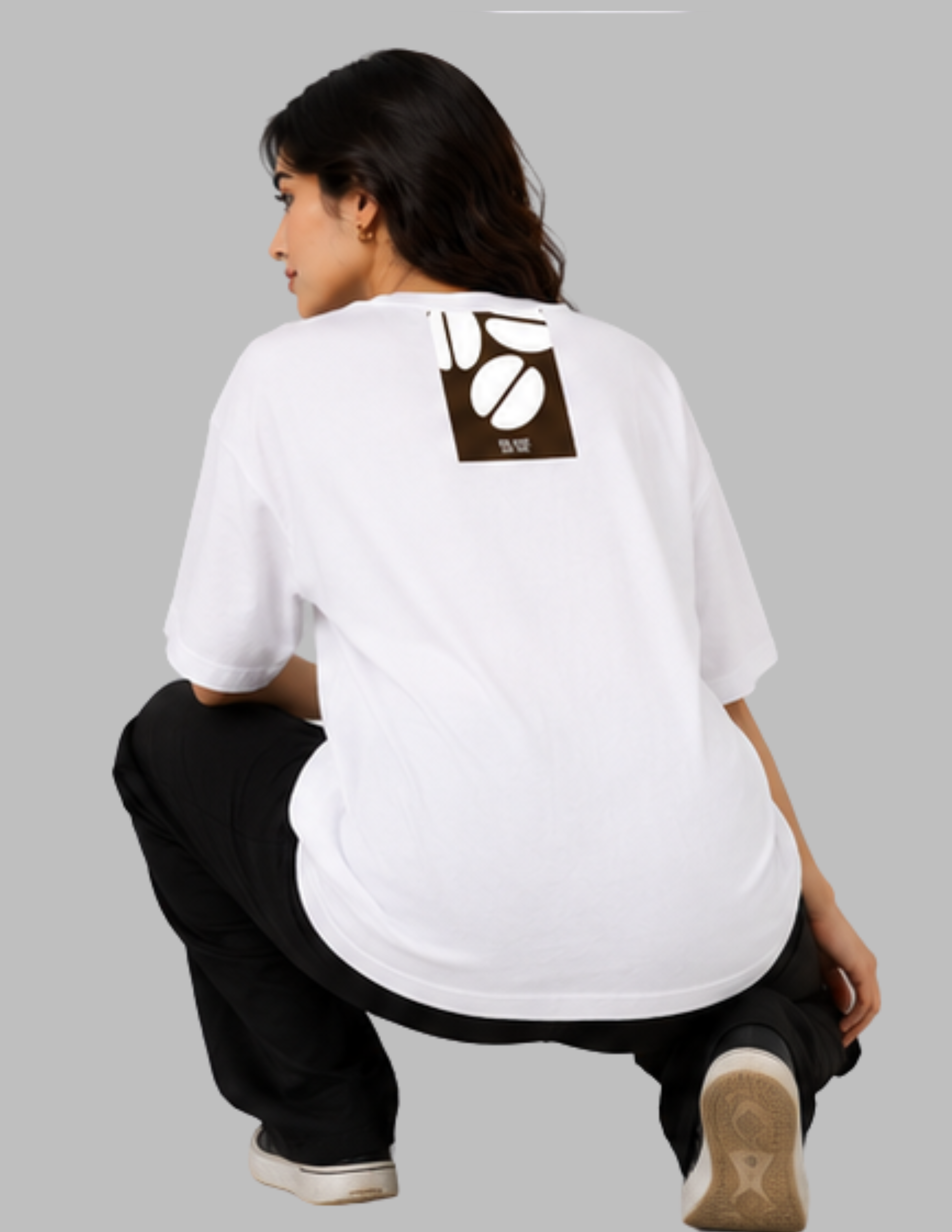A model wearing a white oversized t-shirt with a brown coffee bean graphic on the back, paired with black pants, crouching against a solid gray background.