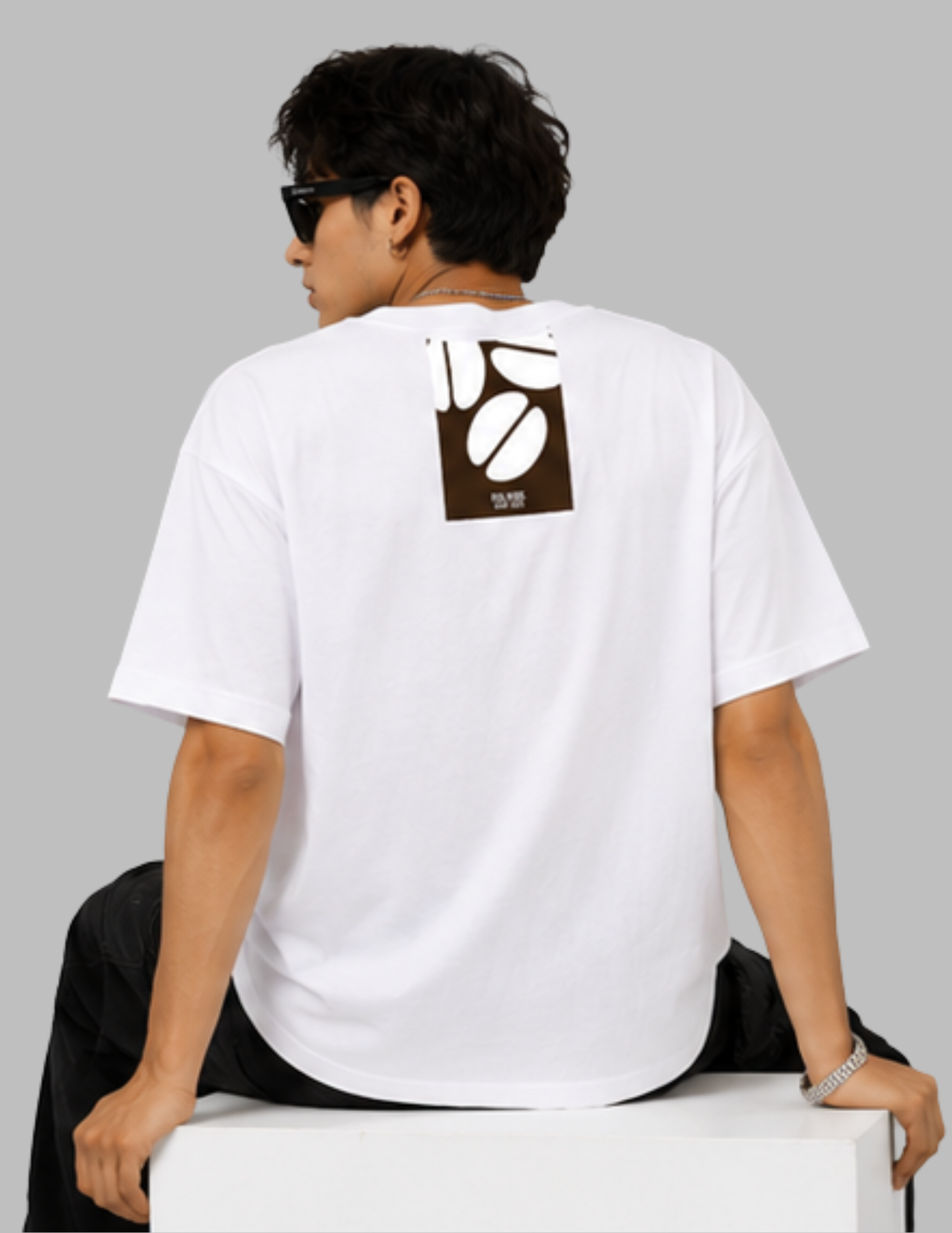 A man wearing a white t-shirt with a brown coffee bean graphic on the back, sitting on a white block against a gray background.