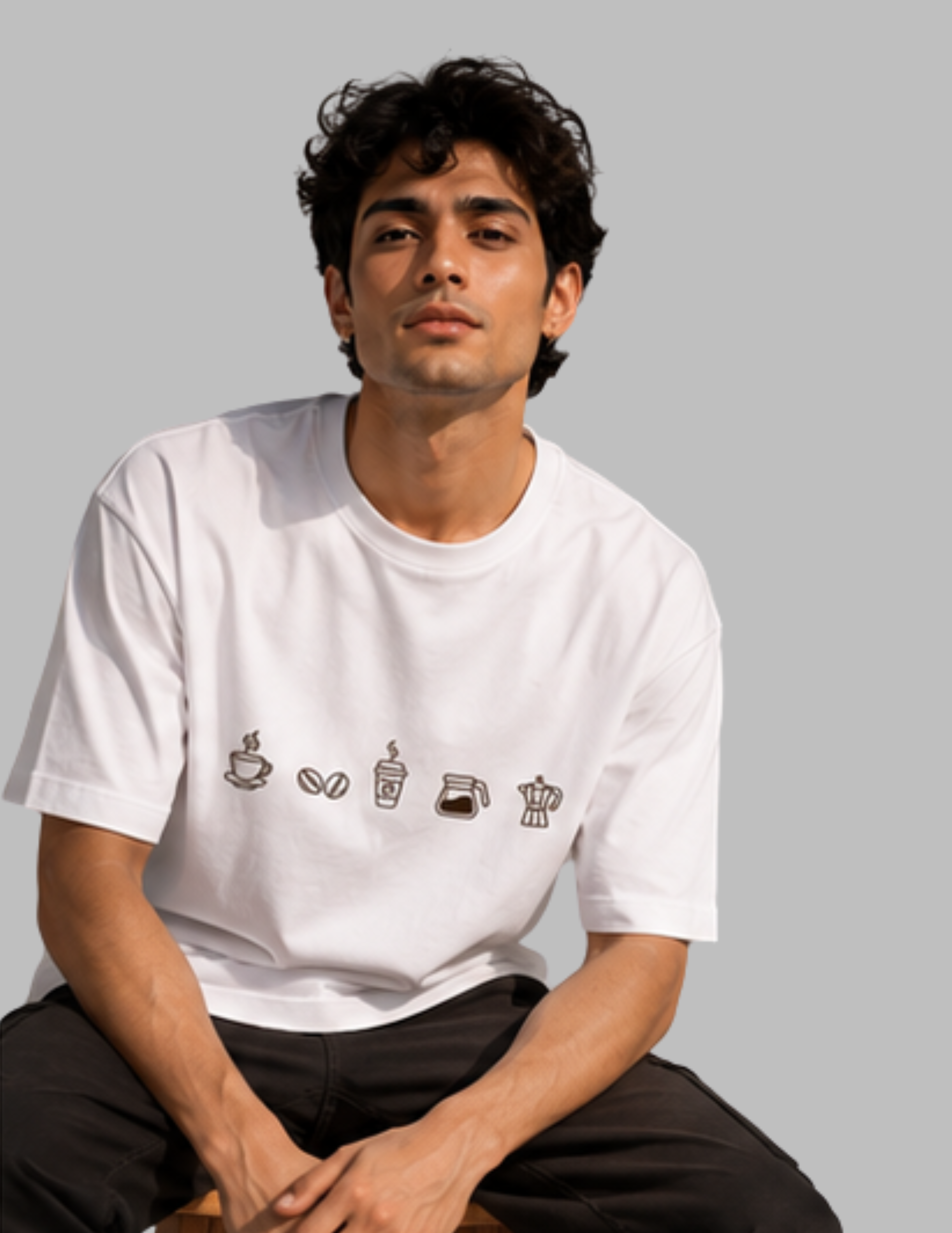 A man wearing a white crewneck t-shirt with a graphic of coffee-related icons on the chest, paired with black pants, set against a solid gray background.