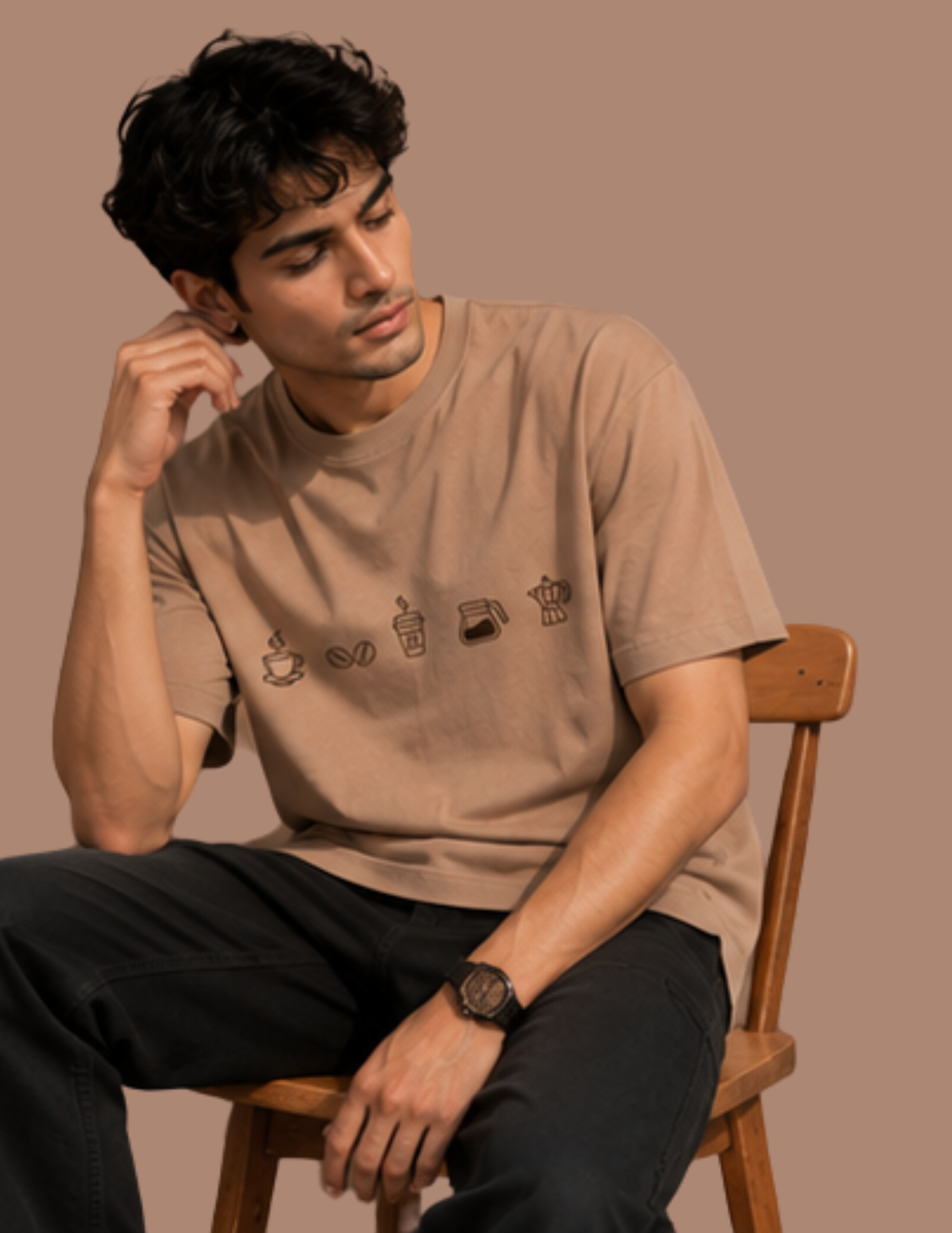 A man wearing a beige graphic t-shirt with coffee-themed illustrations, sitting on a wooden chair against a solid warm brown background.