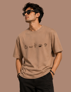 A man wearing a beige crewneck t-shirt with a small graphic of coffee-themed icons on the chest, styled with black sunglasses and black pants against a solid warm brown background.