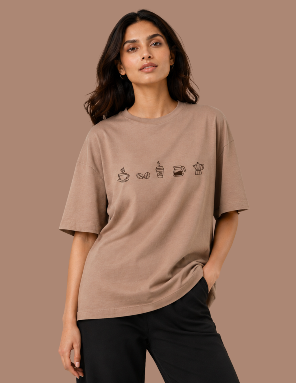 A woman modeling an oversized taupe-brown t-shirt with a row of small coffee-themed line drawings on the chest, paired with black pants, set against a solid taupe background.