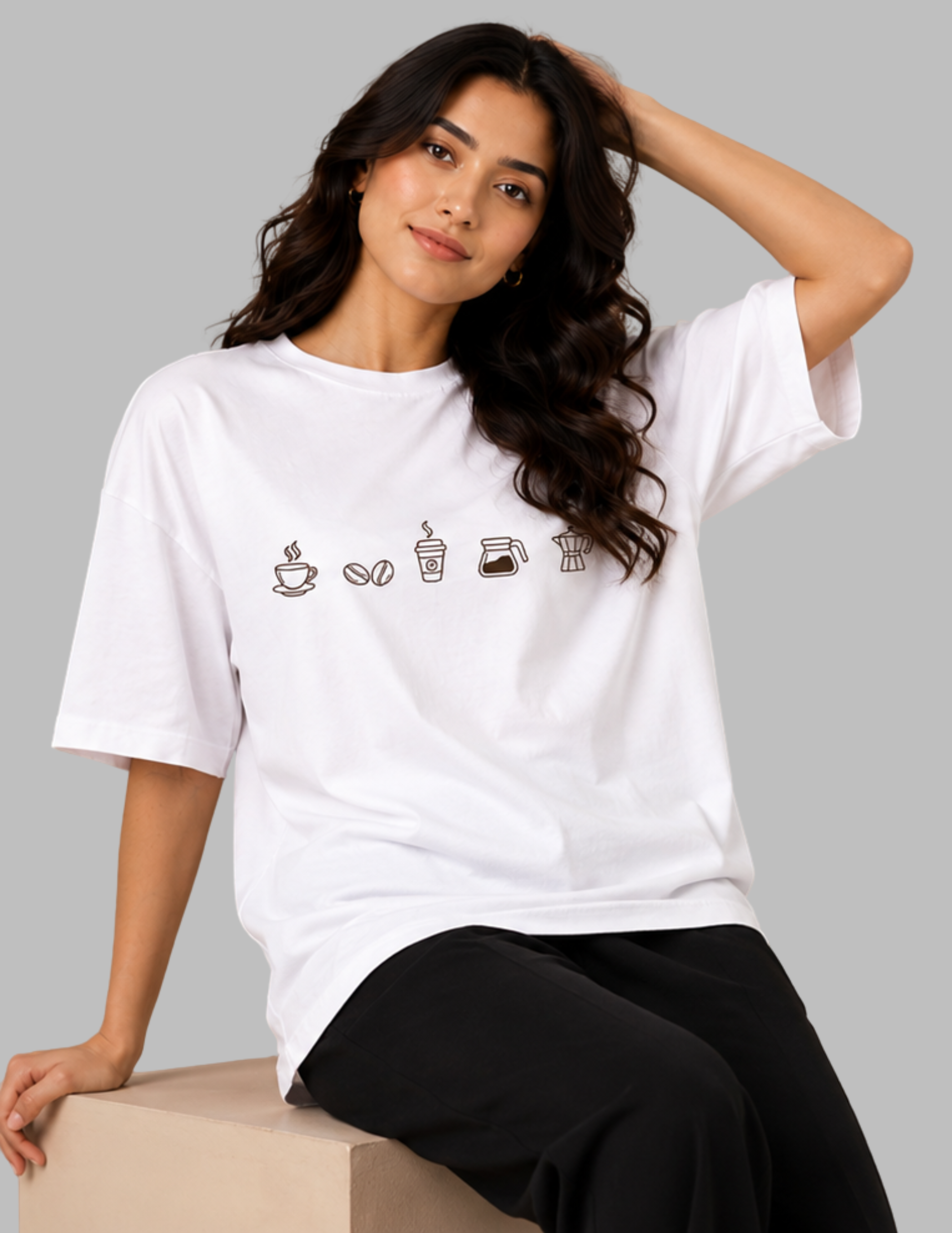 A woman wearing a white short-sleeve t-shirt with a graphic of coffee-themed icons across the chest, paired with black pants, sitting on a beige block against a solid gray background.