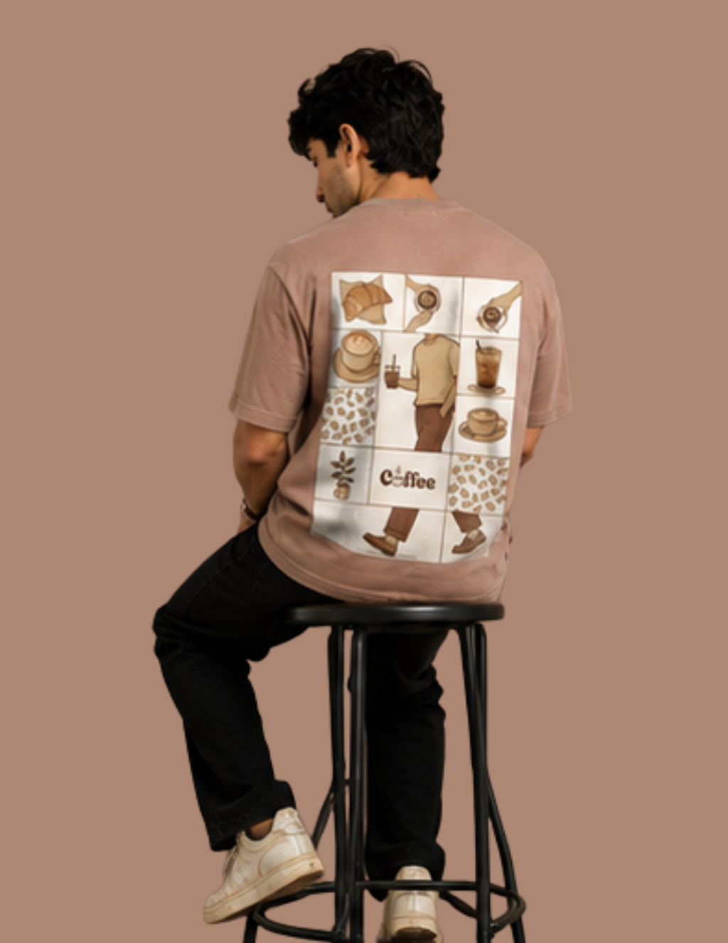 A man wearing a brown graphic t-shirt with a coffee-themed collage print on the back, sitting on a black metal stool against a solid brown background.