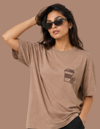 A woman wearing a white oversized T-shirt with a large vintage-style coffee-themed graphic print on the back, seated on a white stool against a solid gray background.