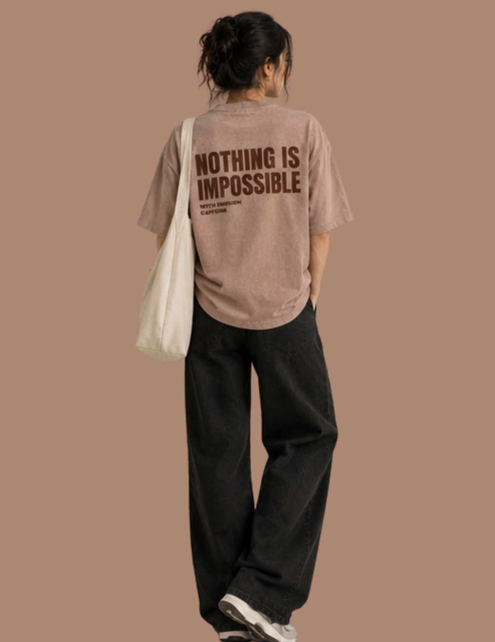 A woman shown from behind wearing a taupe-brown oversized t-shirt with the text 'NOTHING IS IMPOSSIBLE' printed in dark brown on the back, paired with black wide-leg pants and a white canvas shoulder bag, set against a solid taupe background.