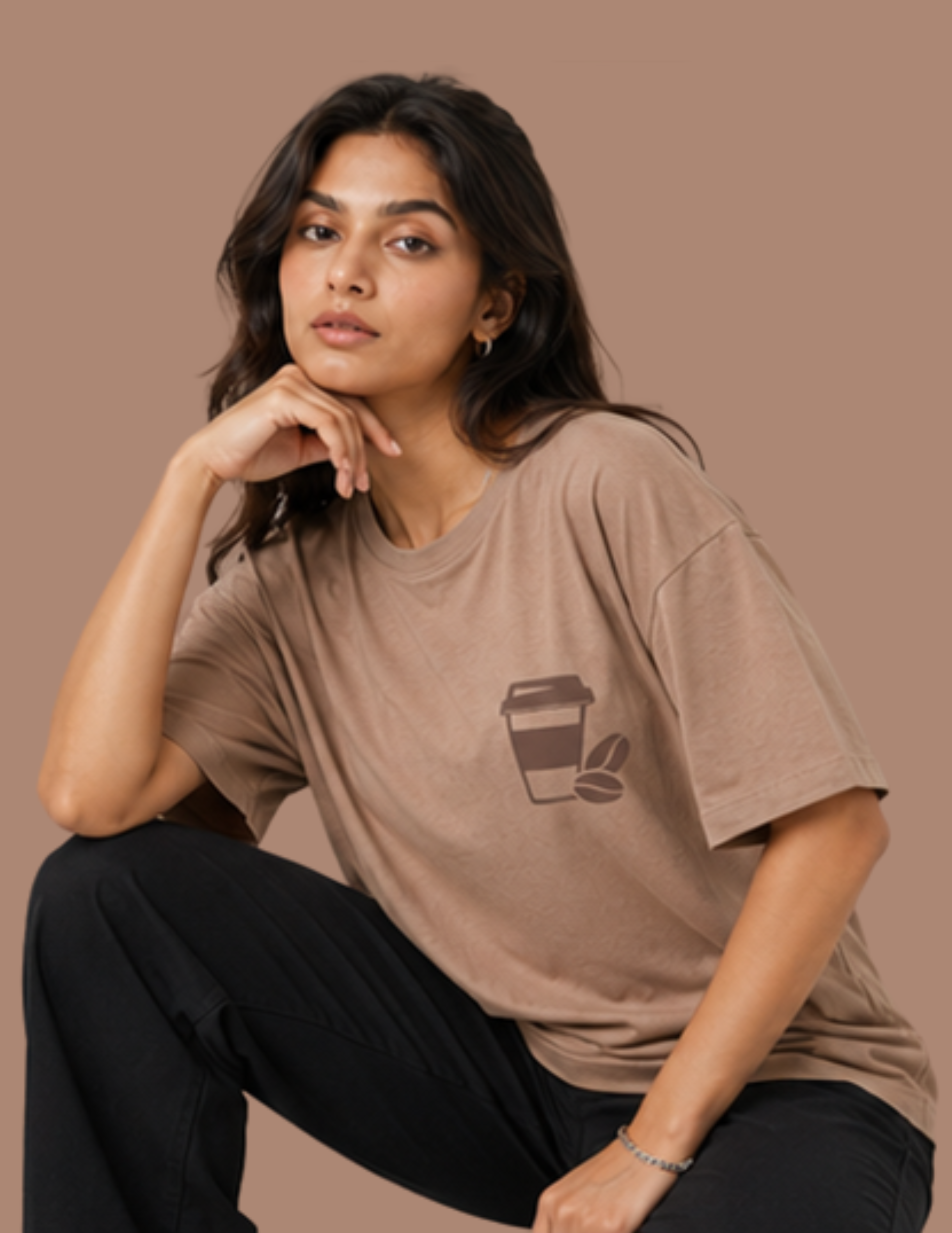 A woman wearing a white oversized T-shirt with a large vintage-style coffee-themed graphic print on the back, seated on a white stool against a solid gray background.