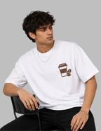 A man wearing a white crewneck t-shirt with a brown coffee cup and coffee bean graphic on the left chest, sitting on a black chair against a solid gray background.