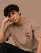 A man wearing a brown short-sleeve t-shirt with a coffee cup graphic on the left chest, posing against a solid brown background.