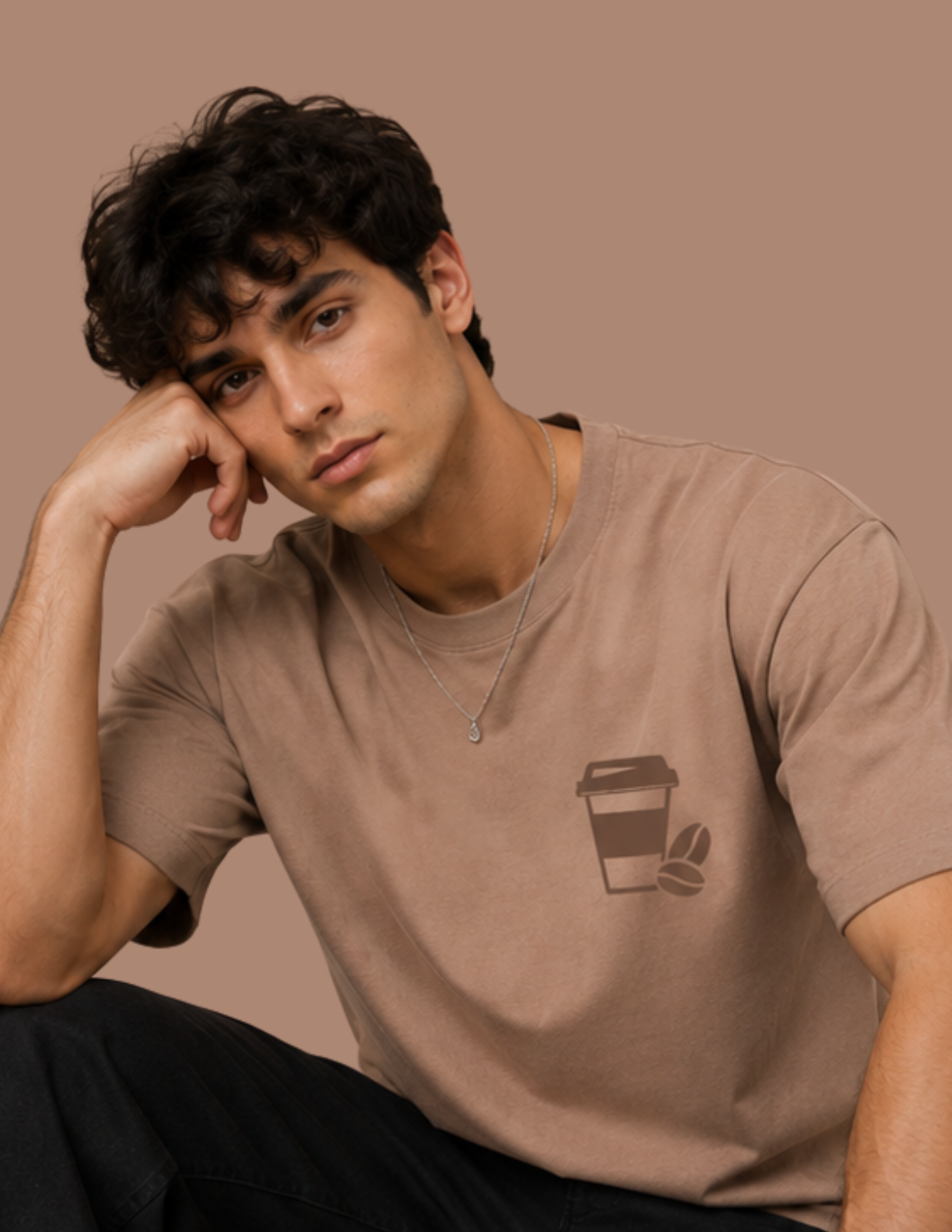 A man wearing a brown short-sleeve t-shirt with a coffee cup graphic on the left chest, posing against a solid brown background.