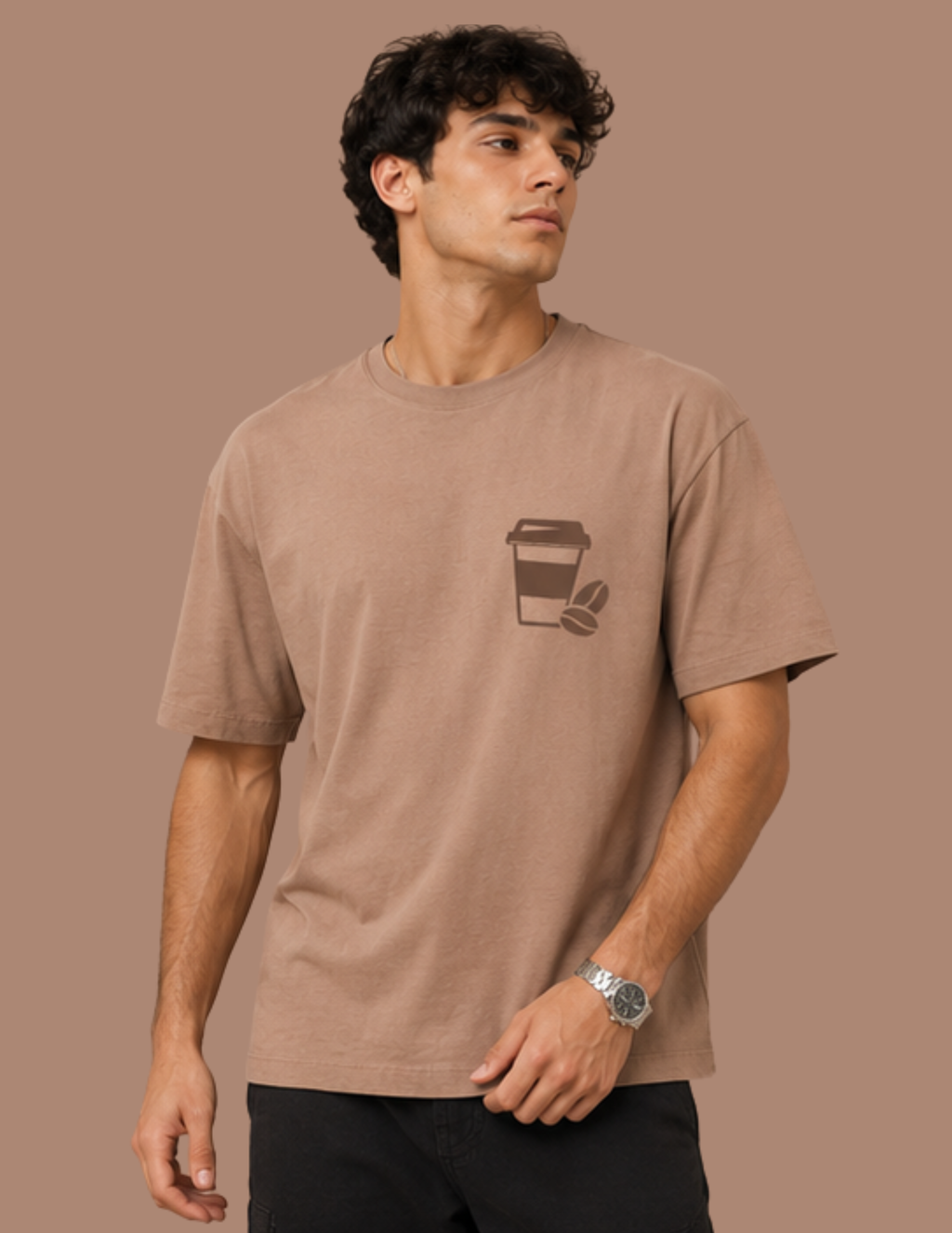 A man wearing a light brown crewneck t-shirt with a dark brown coffee cup graphic on the left chest, posed against a solid brown background.