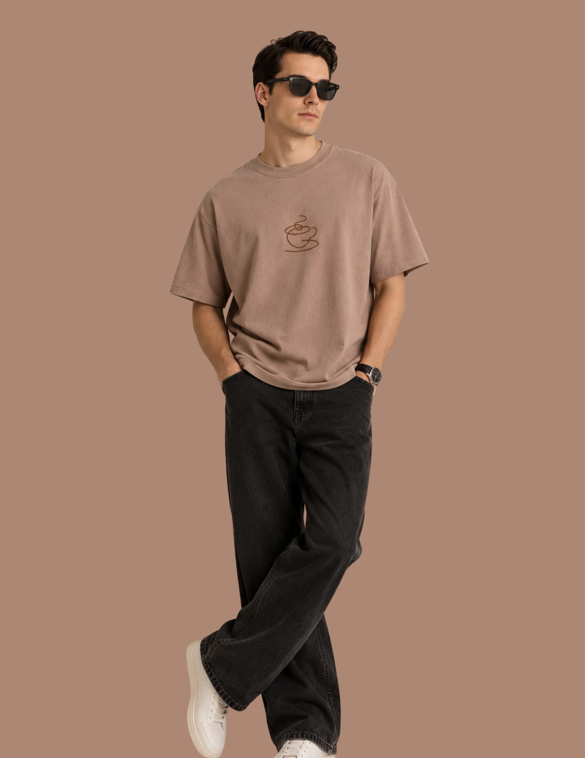 A man wearing a light brown crewneck t-shirt with a small coffee cup graphic on the chest, paired with black jeans and white sneakers, standing against a solid brown background.