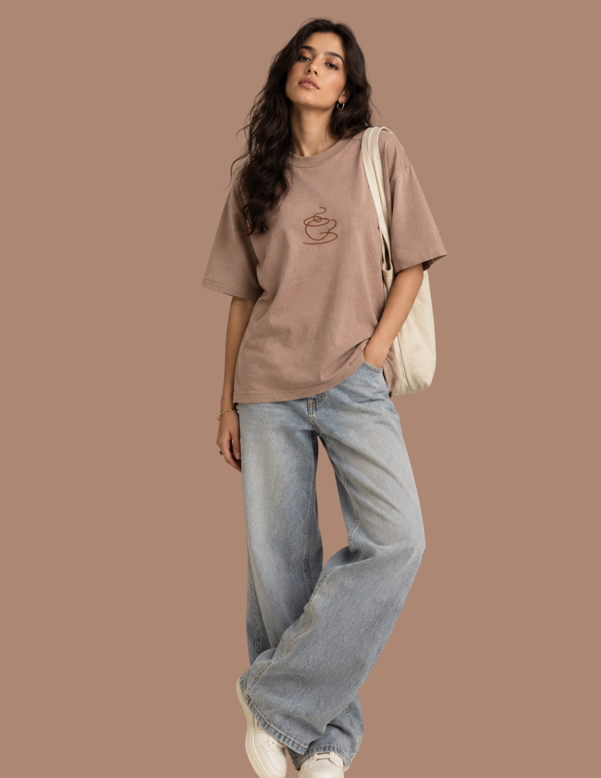 A woman modeling an oversized taupe-brown t-shirt with a small coffee cup graphic on the chest, paired with light-wash wide-leg jeans, white sneakers, and a cream canvas shoulder bag, set against a solid warm brown background.