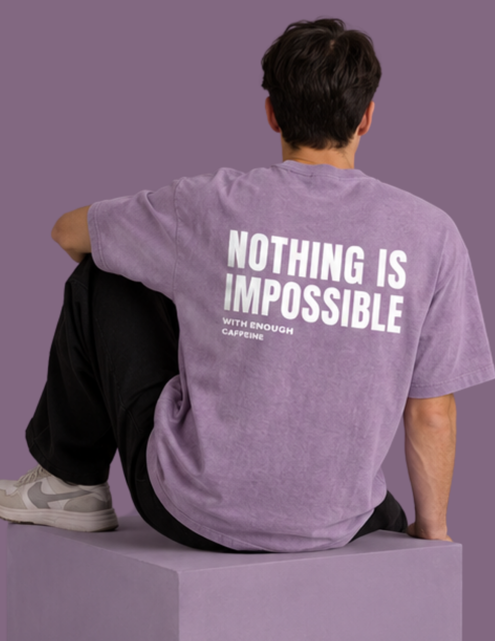 A man sits on a lavender block, viewed from behind, wearing a heathered lavender t-shirt with bold white text reading 'NOTHING IS IMPOSSIBLE WITH ENOUGH CAFFEINE' on the back. The background is a solid lavender-purple color.
