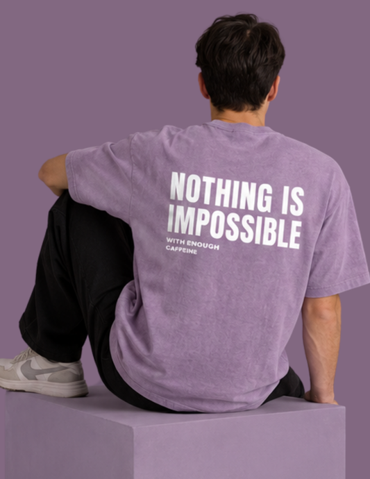 A man sits on a lavender block, viewed from behind, wearing a heathered lavender t-shirt with bold white text reading 'NOTHING IS IMPOSSIBLE WITH ENOUGH CAFFEINE' on the back. The background is a solid lavender-purple color.