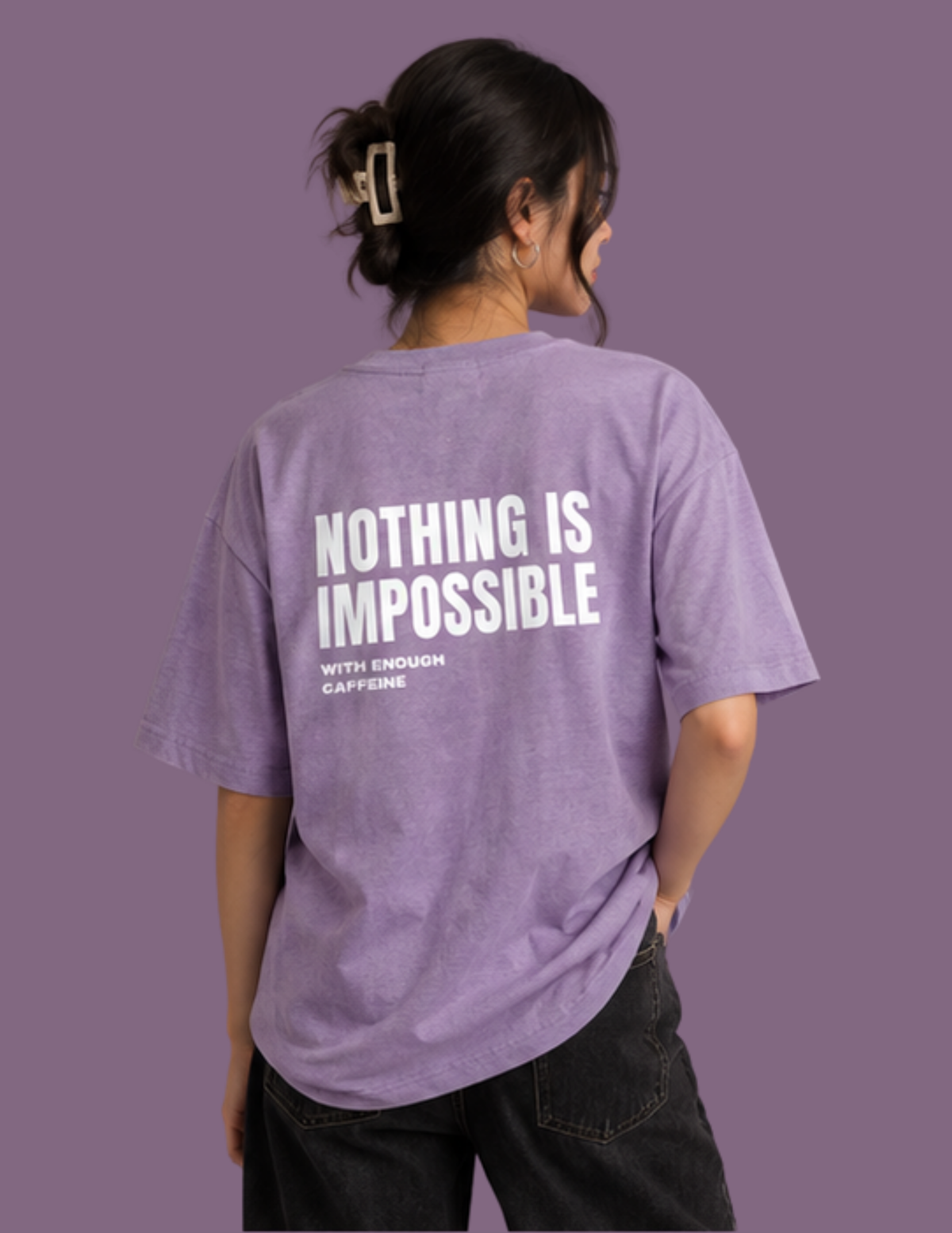 A woman shown from behind wearing a lavender-purple graphic t-shirt with the text 'NOTHING IS IMPOSSIBLE WITH ENOUGH CAFFEINE' printed in white on the back, paired with black jeans, set against a solid purple background.
