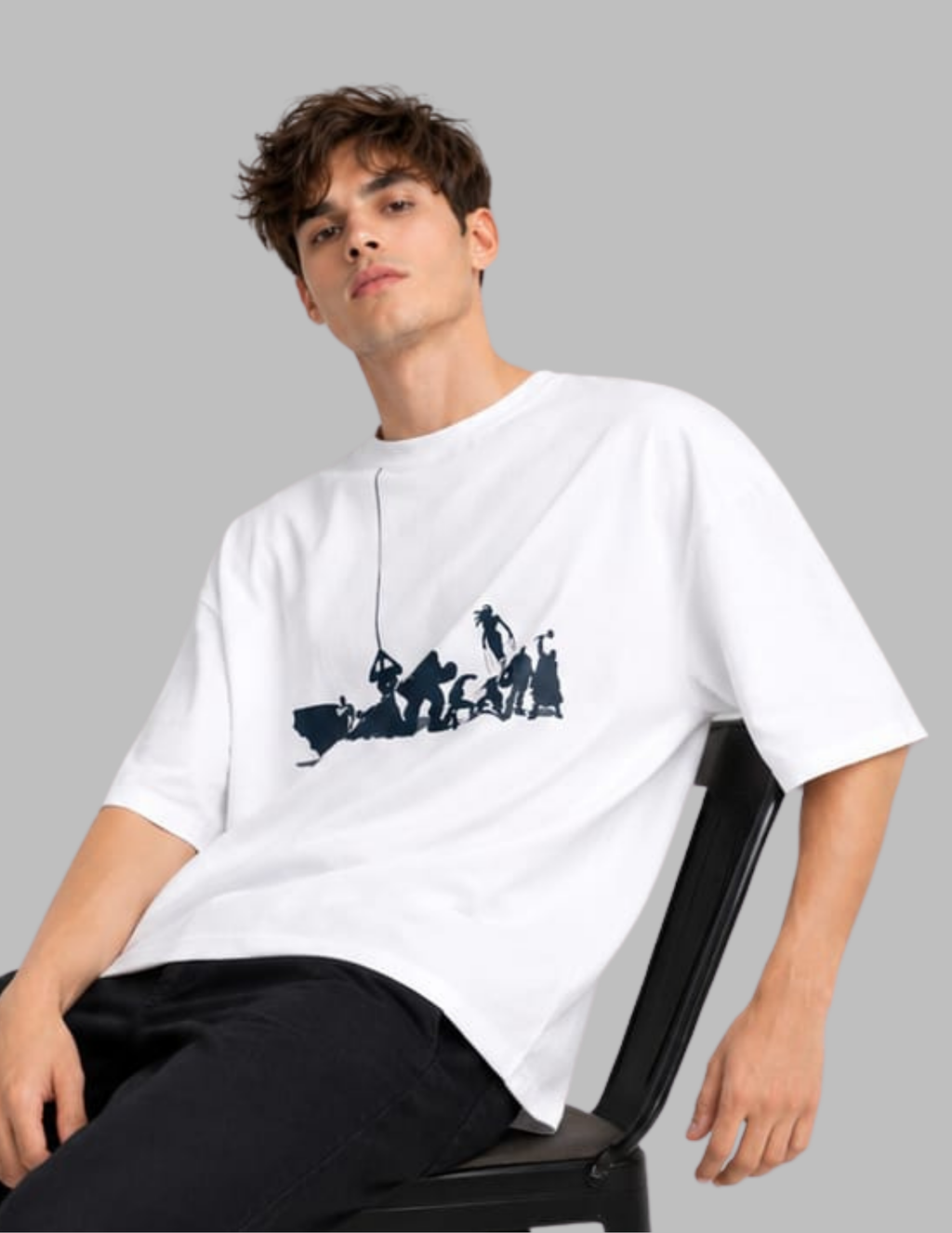 A man wearing a white oversized t-shirt with a dark blue graphic print of figures on a boat, seated on a black chair against a solid gray background.