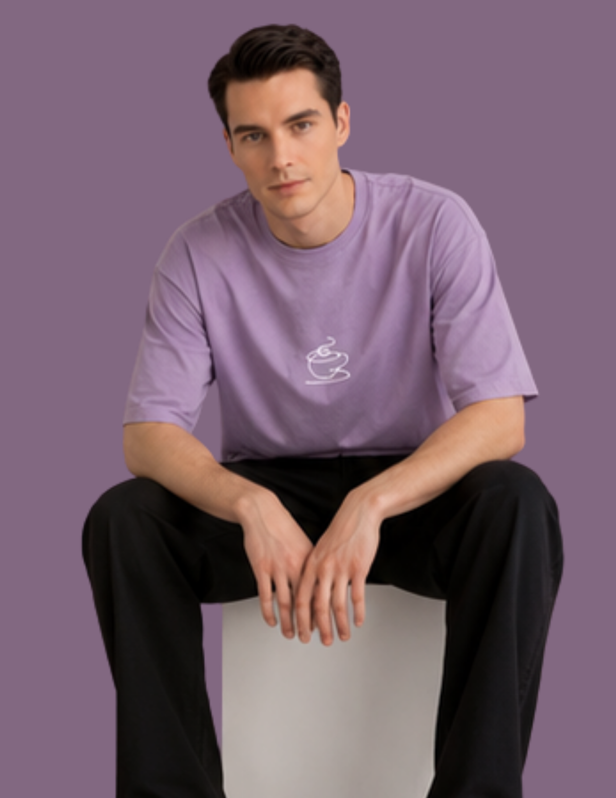 A man wearing a lavender purple t-shirt with a small white coffee cup graphic on the chest, paired with black pants, sitting on a white block against a solid purple background.