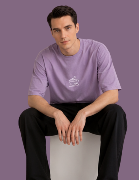 A man wearing a lavender purple t-shirt with a small white coffee cup graphic on the chest, paired with black pants, sitting on a white block against a solid purple background.
