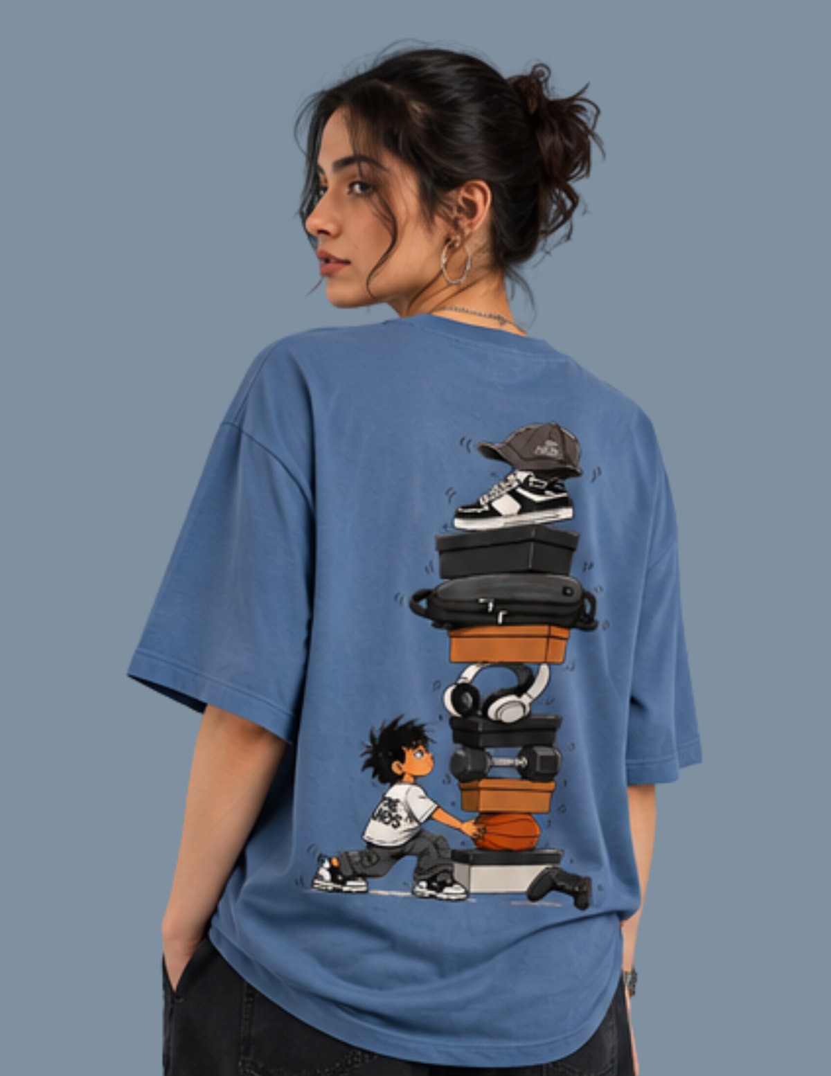 A woman shown from the back wearing a blue oversized T-shirt with a large graphic print of a child pushing a stack of shoes, headphones, and cases, against a solid slate blue background.