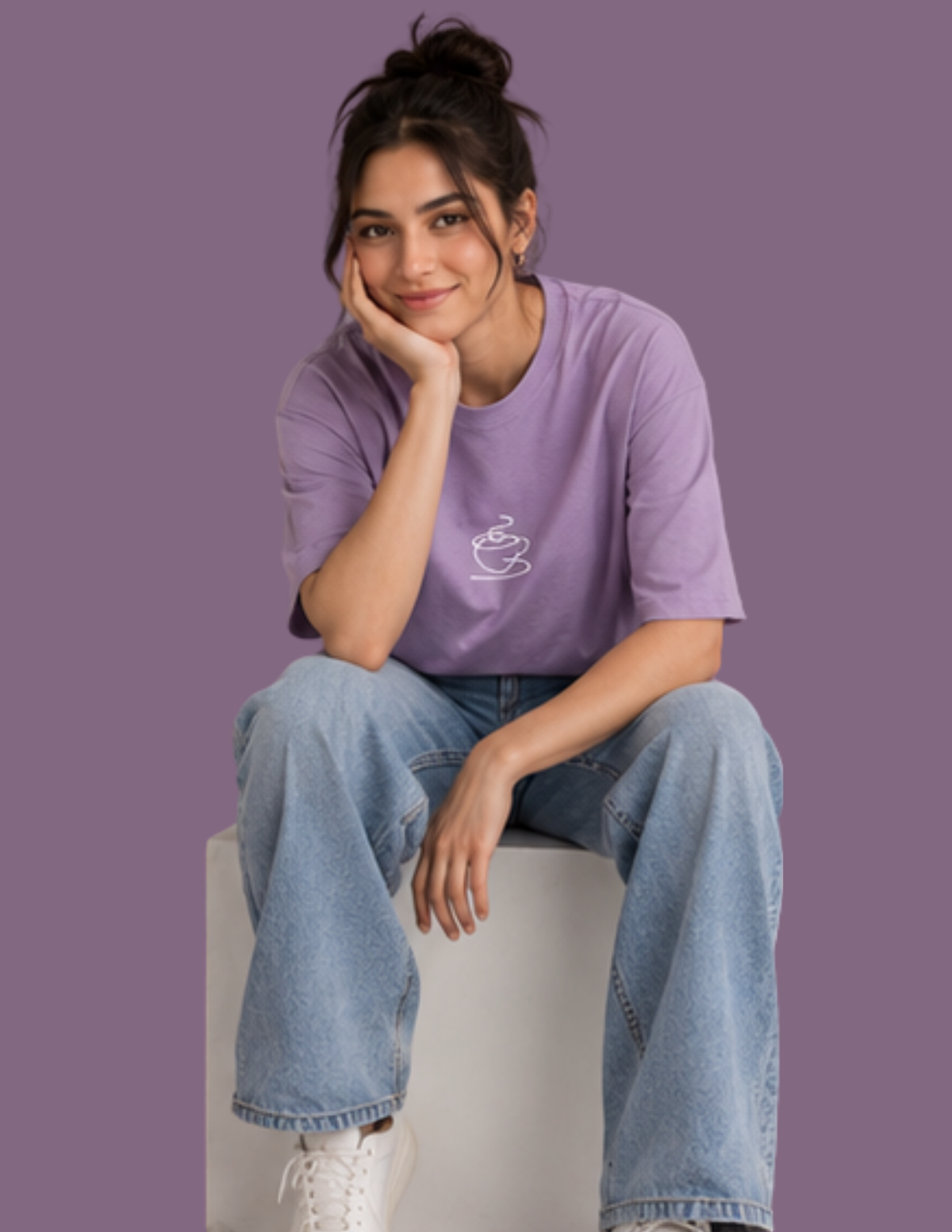 A woman sitting on a white block wearing a lavender purple t-shirt with a small white coffee cup graphic on the chest, paired with light blue jeans and white sneakers, against a solid purple background.