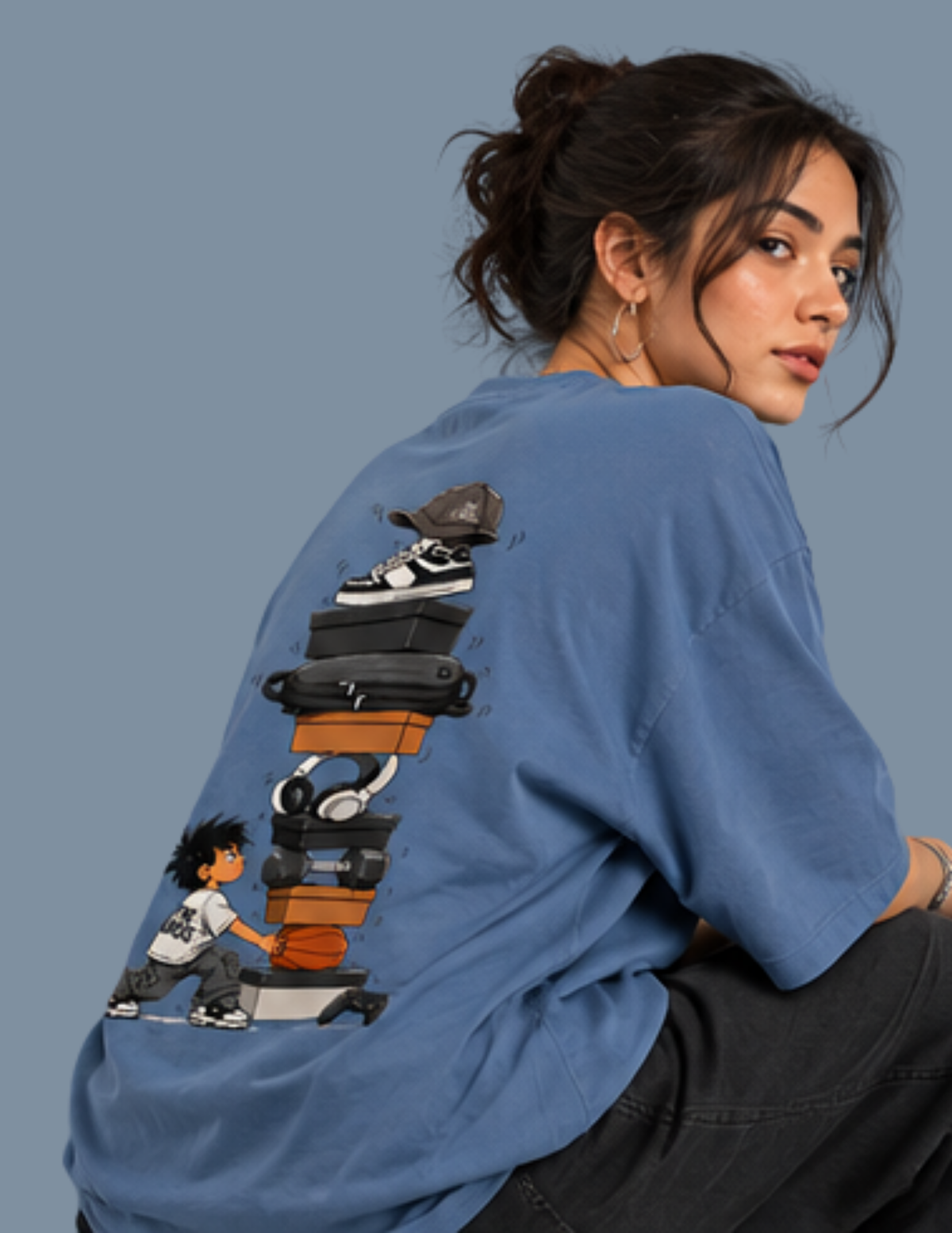 A woman wearing a blue oversized t-shirt with a large graphic print on the back, featuring a cartoon character and stacked sneakers, against a solid blue-gray background.