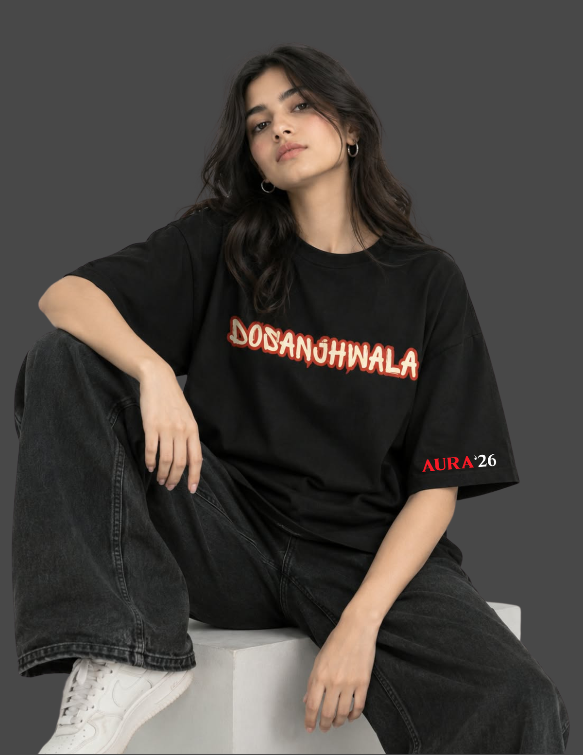 A woman sits on a white block wearing a black oversized graphic T-shirt with 'DOSANGHWALA' across the chest in red and orange outlined lettering, plus 'AURA26' on the sleeve, paired with dark jeans and white sneakers against a dark gray background.