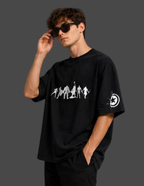 A man wearing a black oversized t-shirt with a white graphic print of figures on the chest and a circular logo on the sleeve, paired with black pants and sunglasses, set against a dark gray background.