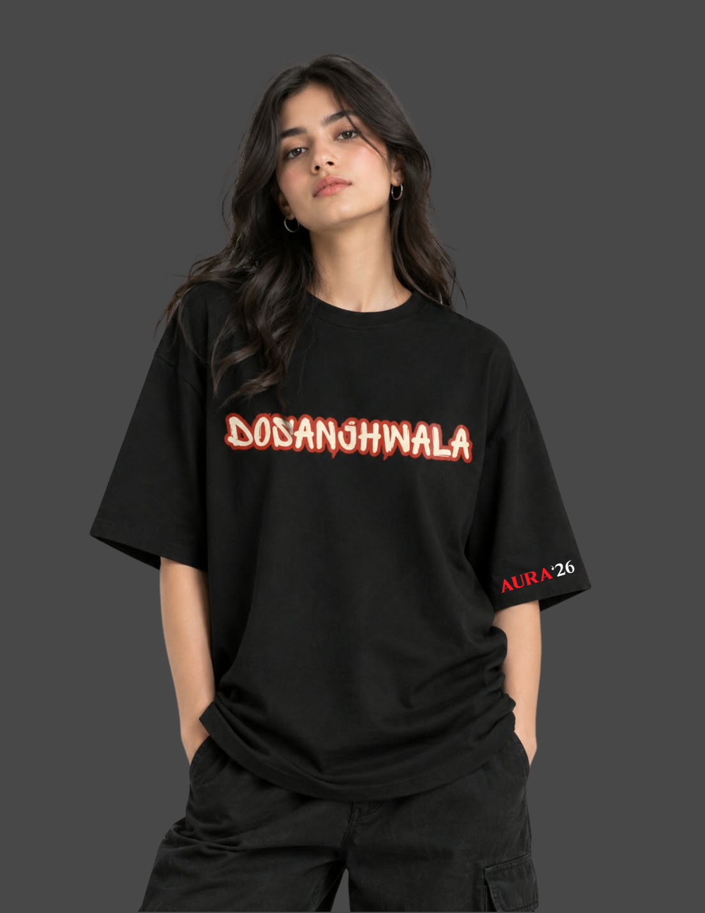 A woman wearing a black oversized graphic T-shirt with 'DONANGHWALA' printed across the chest in red and orange lettering, and 'AURA26' on the right sleeve, against a dark gray background.