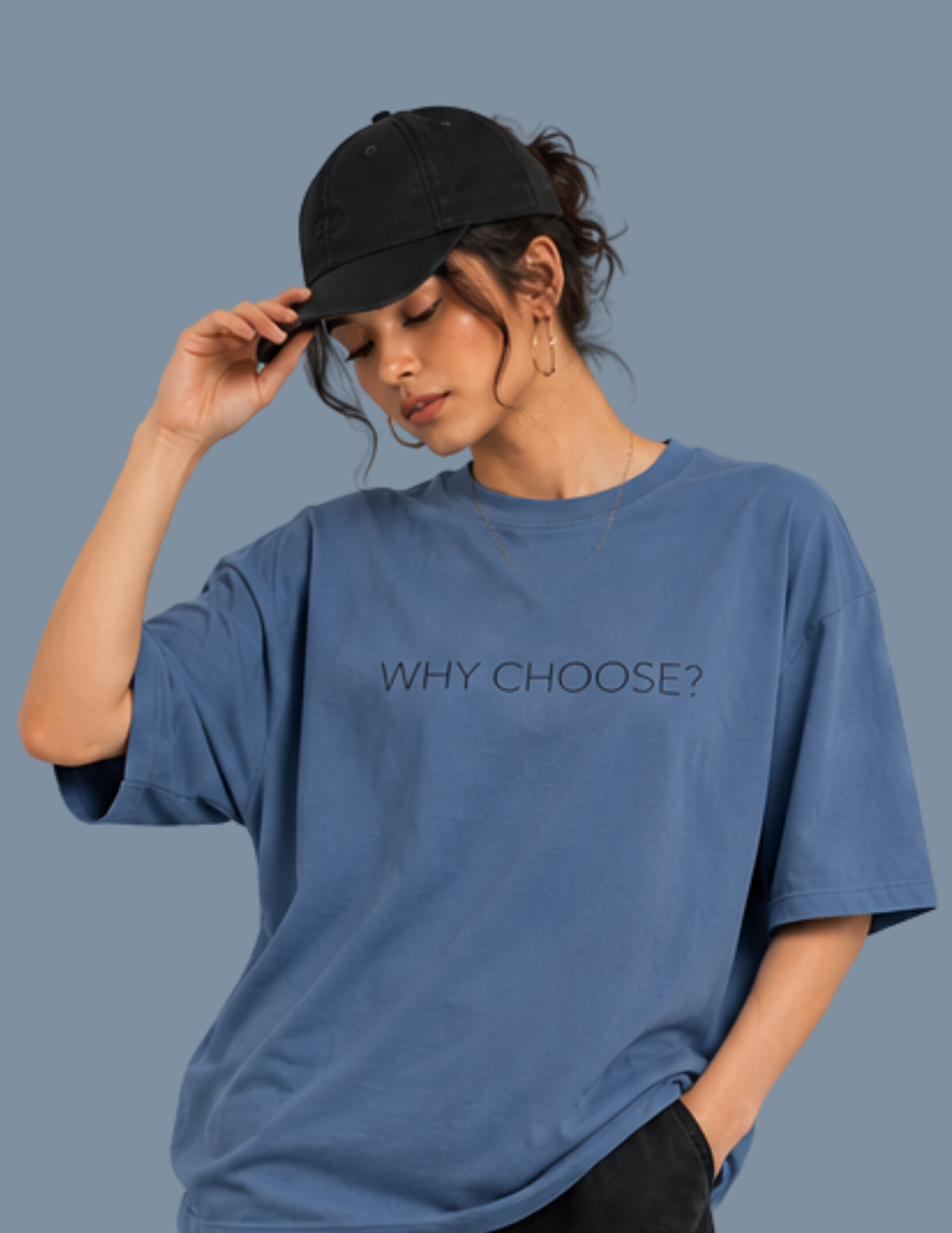A woman wearing a blue oversized t-shirt with the text 'WHY CHOOSE?' printed on the chest, paired with a black baseball cap, set against a solid slate blue background.