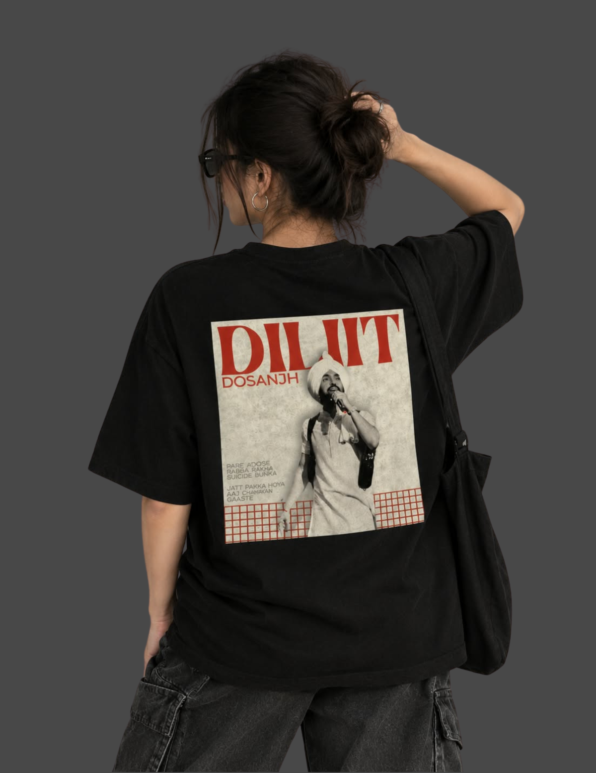 A woman shown from behind wearing a black oversized T-shirt with a large back graphic featuring the text 'DILJIT DOSANJH' and a monochrome portrait print, against a dark gray studio background.