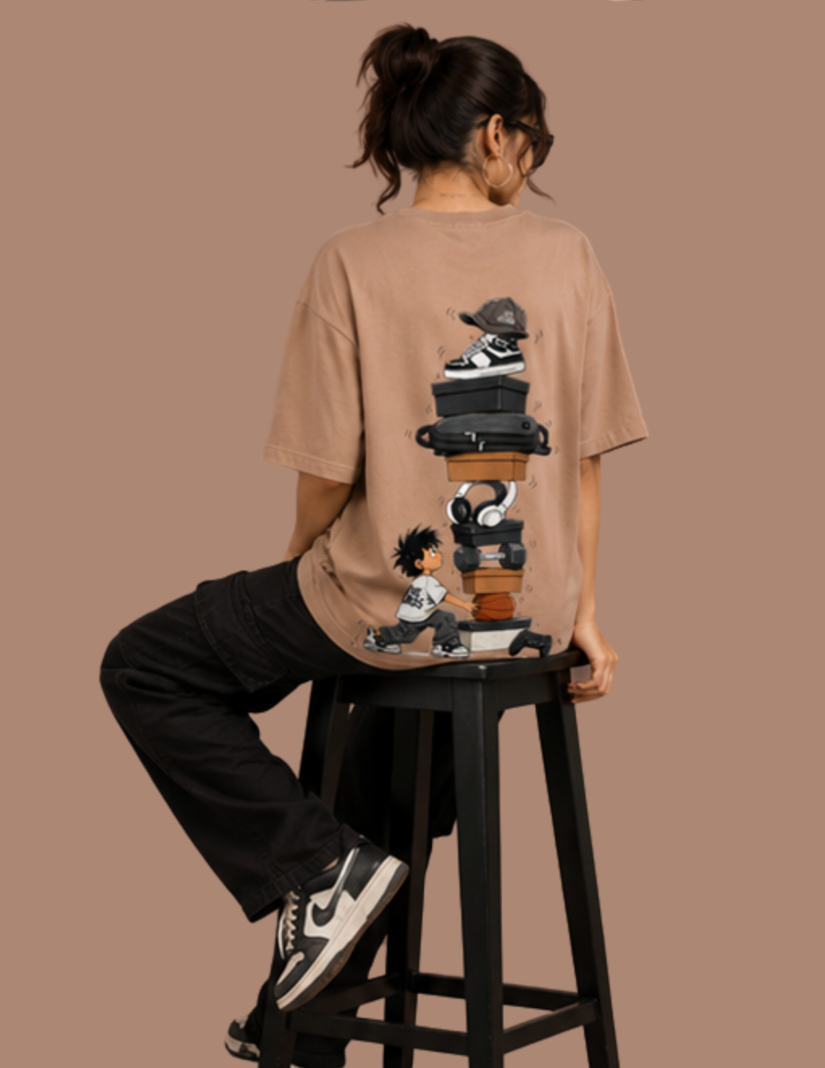 A model wearing a beige graphic t-shirt, sitting on a black stool and viewed from behind. The t-shirt features a detailed graphic of stacked sneakers and a cartoon character on the back.