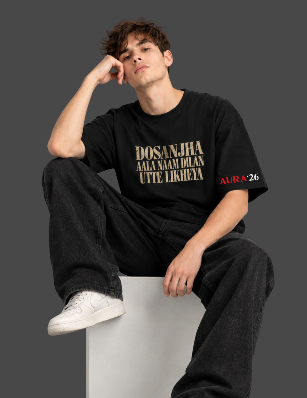 A man wearing a black graphic t-shirt with gold and red text, paired with black jeans and white sneakers, sitting on a white block against a dark gray background.